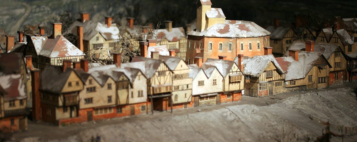 A model box of a wooden circular theatre surrounded by smaller Tudor buildings, along the bank of a snowy and frozen River Thames.