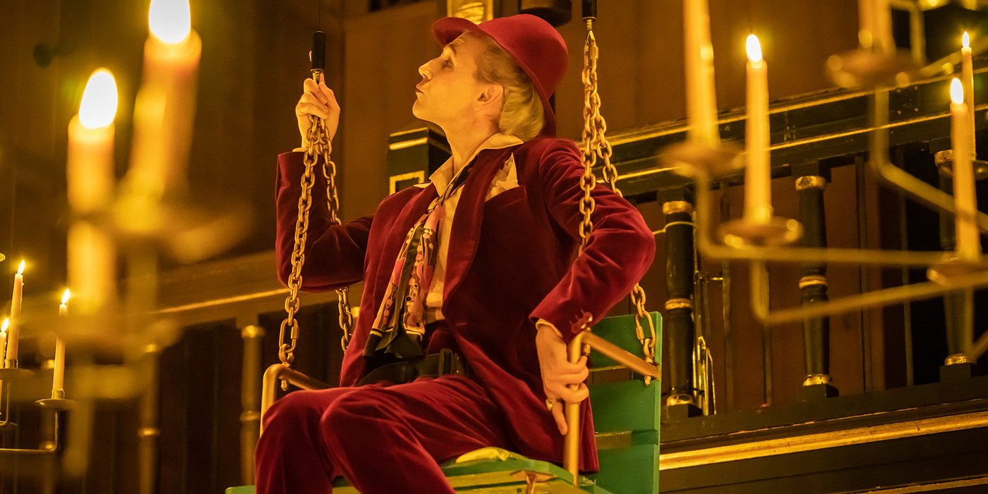 An actor wearing a deep red velvet suit sits on a green swing surrounded by lit candles.