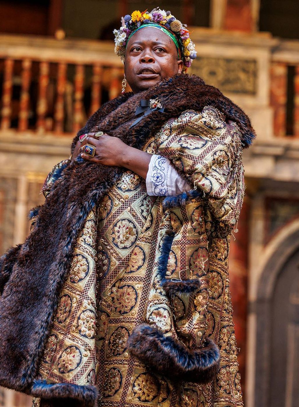 An actor wearing a long golden jacket lined with fur, and a flower crown, stands on stage.