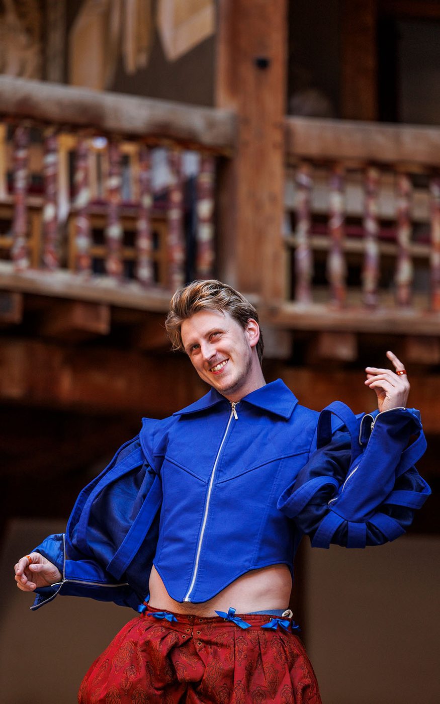 An actor wearing a bright blue zip up doublet stands on the Globe stage.