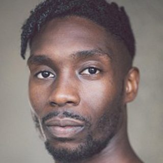 A man with short black dreadlocks looks into the camera