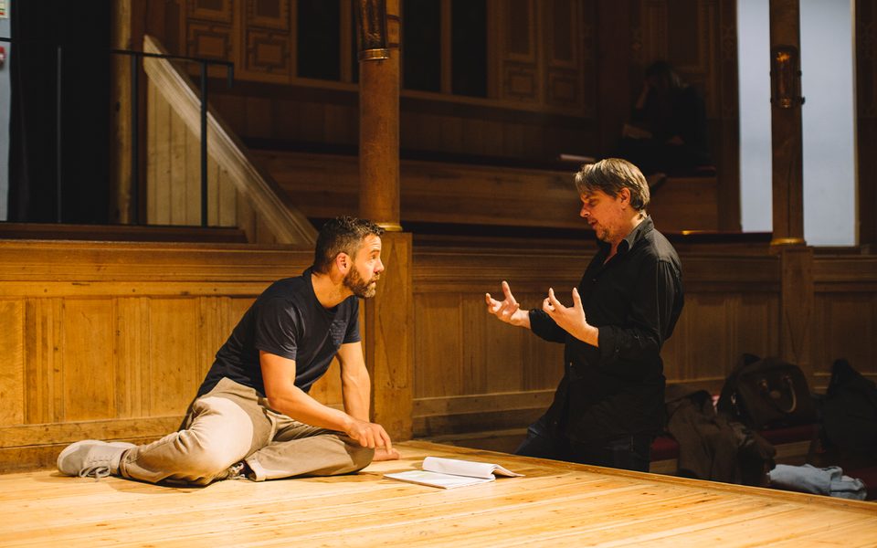 A director dressed all in black talks to an actor who is kneeling on stage with a script in front of them