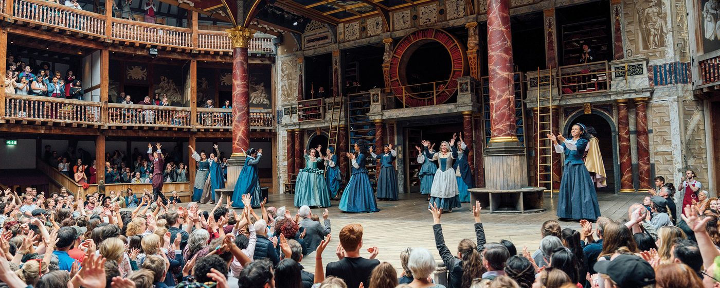 The cast of 'Emilia' gather on stage with their arms in the air, the yard audience also have their arms in the air