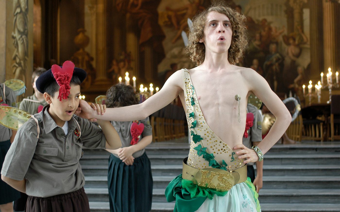 An actor dressed in a gold leotard and green skirt has his hand around the neck of a young actor