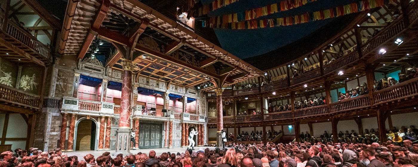 A full audiences watches an actor on stage at nighttime in a circular wooden theatre.