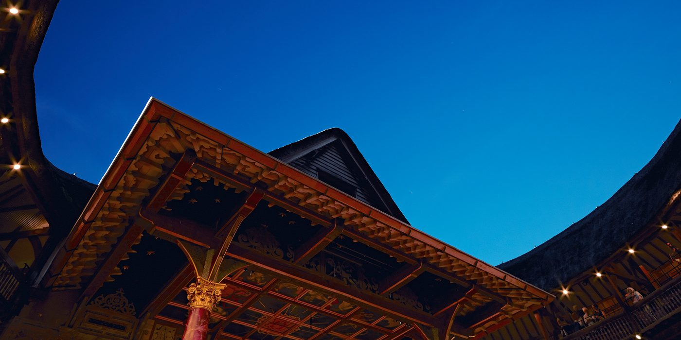 A bright blue evening sky above a circular timber theatre, with pointed thatched roof.