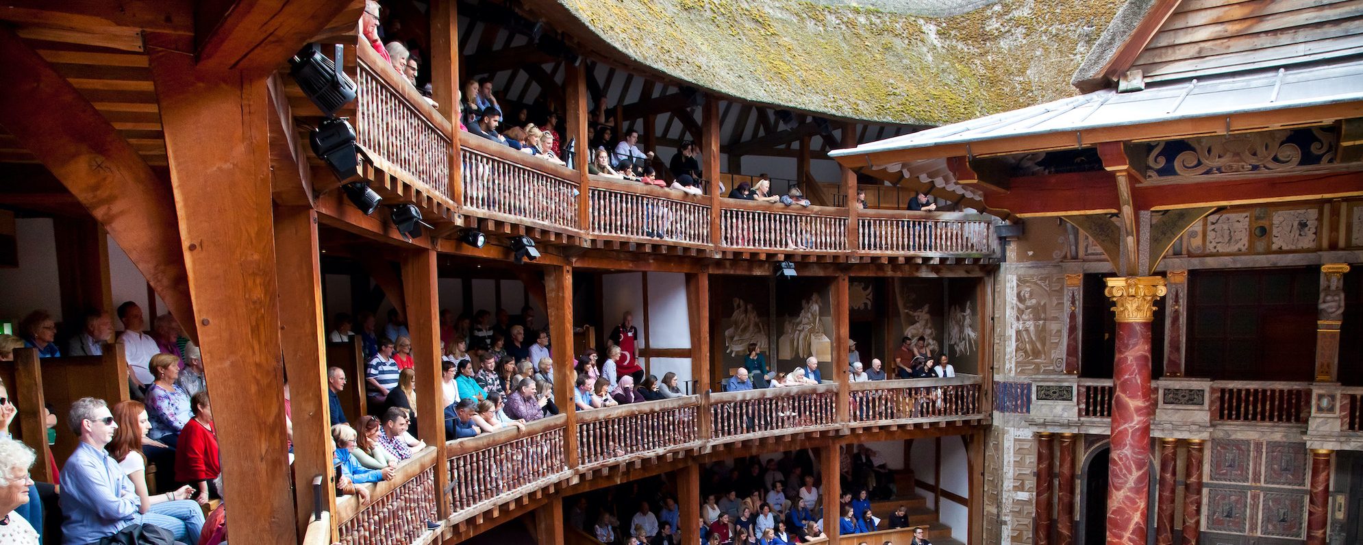 Audience members sit on an outdoor balcony watching a play in the Globe Theatre