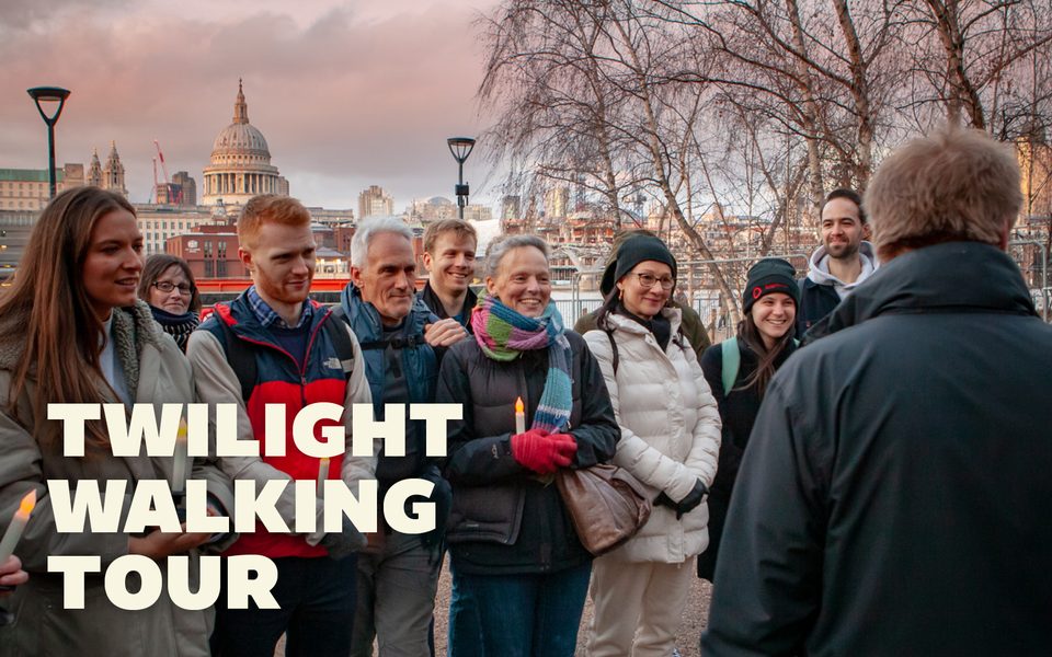 A group of people dressed in coats and scarves stand in a circle listening to a man speak. St Paul's Cathedral is visible in the background.