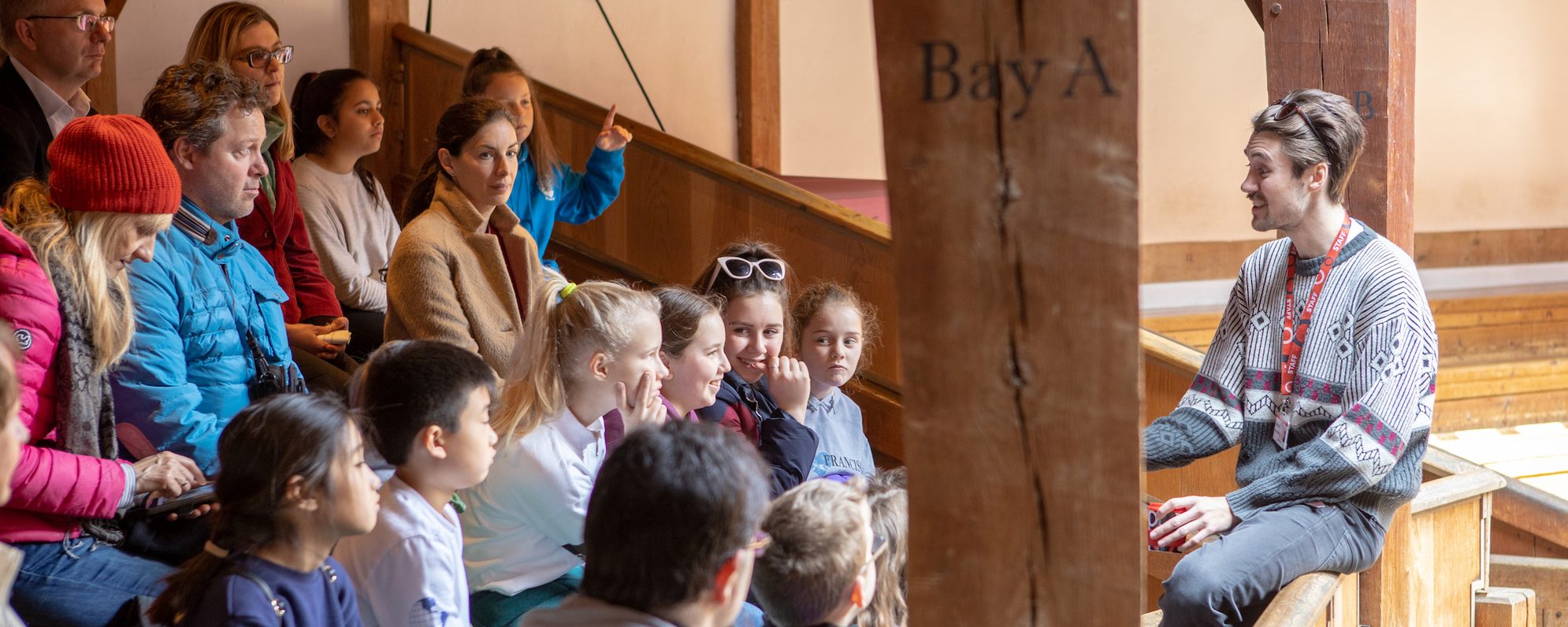 A guide talks to a group who are sitting on wooden benches in a theatre