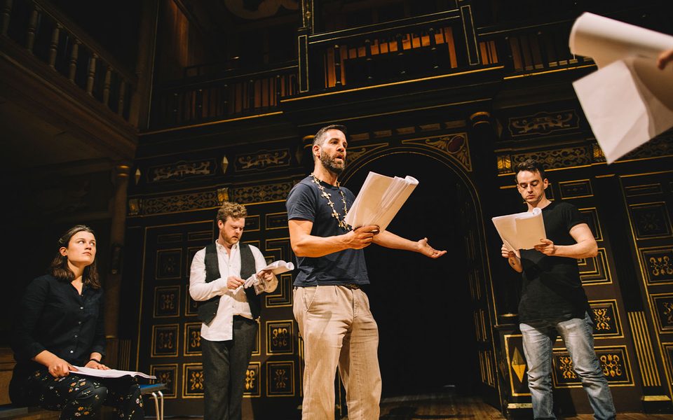 A group of actors performing with scripts in hand. The actors are stood in a circle around one actor who is wearing a golden necklace