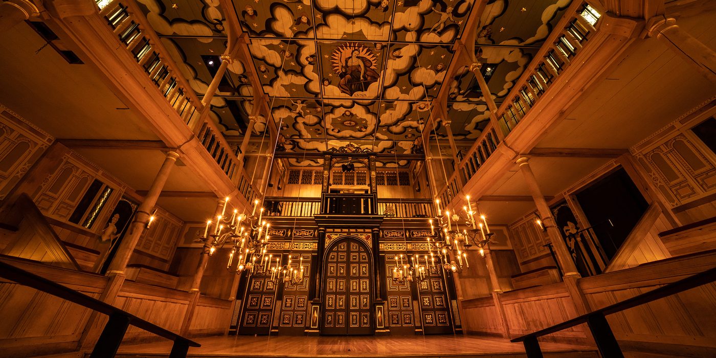 A wide angled shot of the Sam Wanamaker Playhouse with its ornate painting ceiling depicting the Goddess Luna and clouds, hanging candelabra, and panelled painted doors.