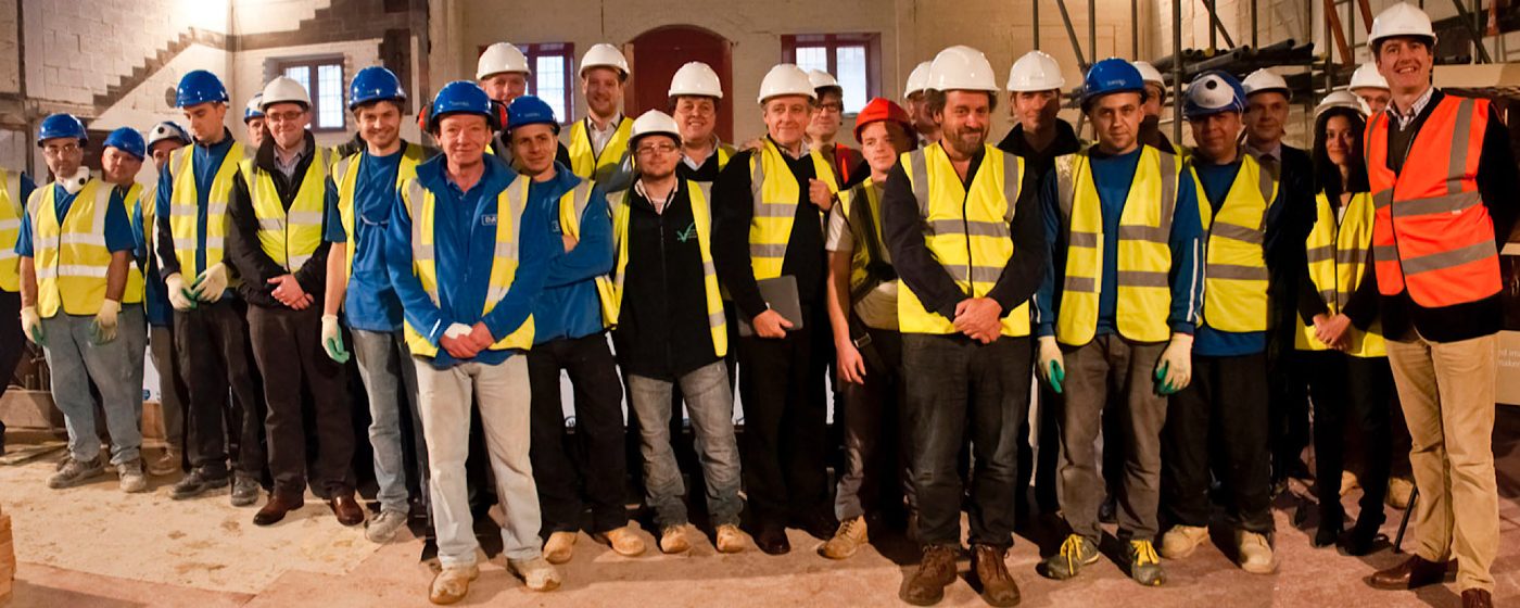 A large group of people in high visibility jackets and hard hats stand on an indoor construction site