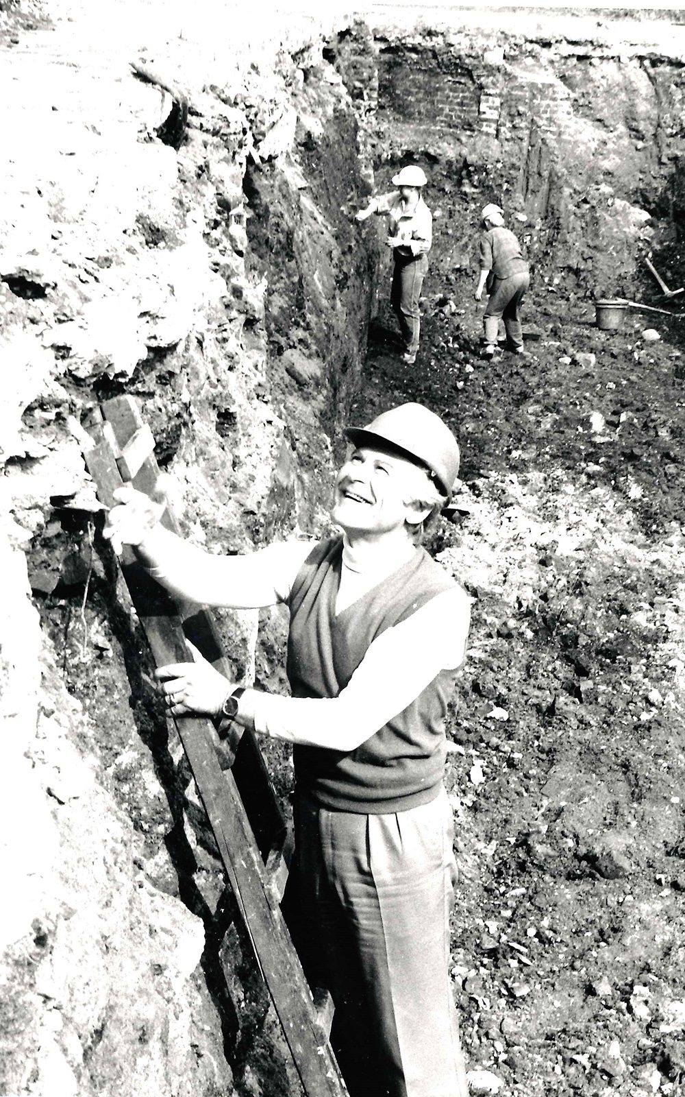 A man stands on a ladder in a ditch of mud and dirt, wearing a hard hat