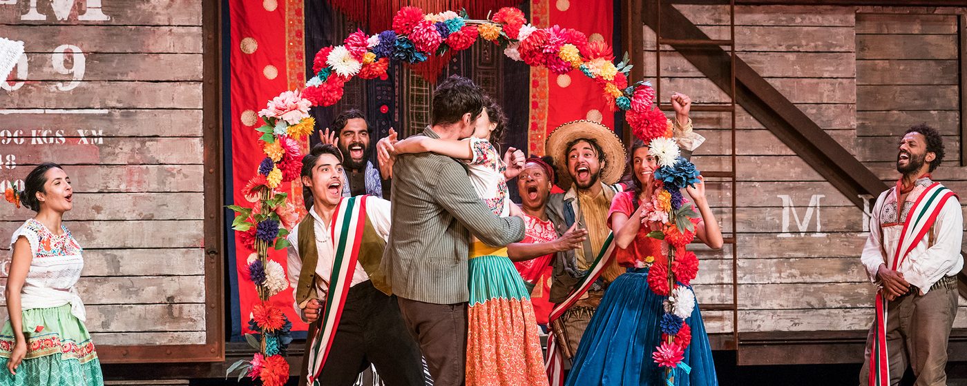 Beatrice and Benedick embrace and kiss surrounded by onlookers. The stage is full of brightly coloured decorations and costumes.