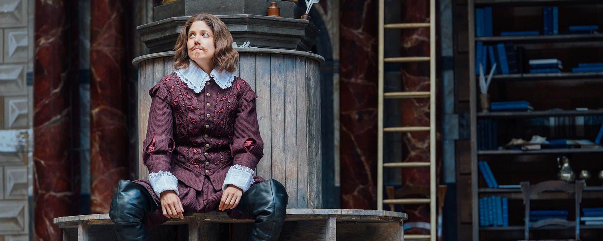 An actor playing William Shakespeare sits by a pillar on the Globe stage