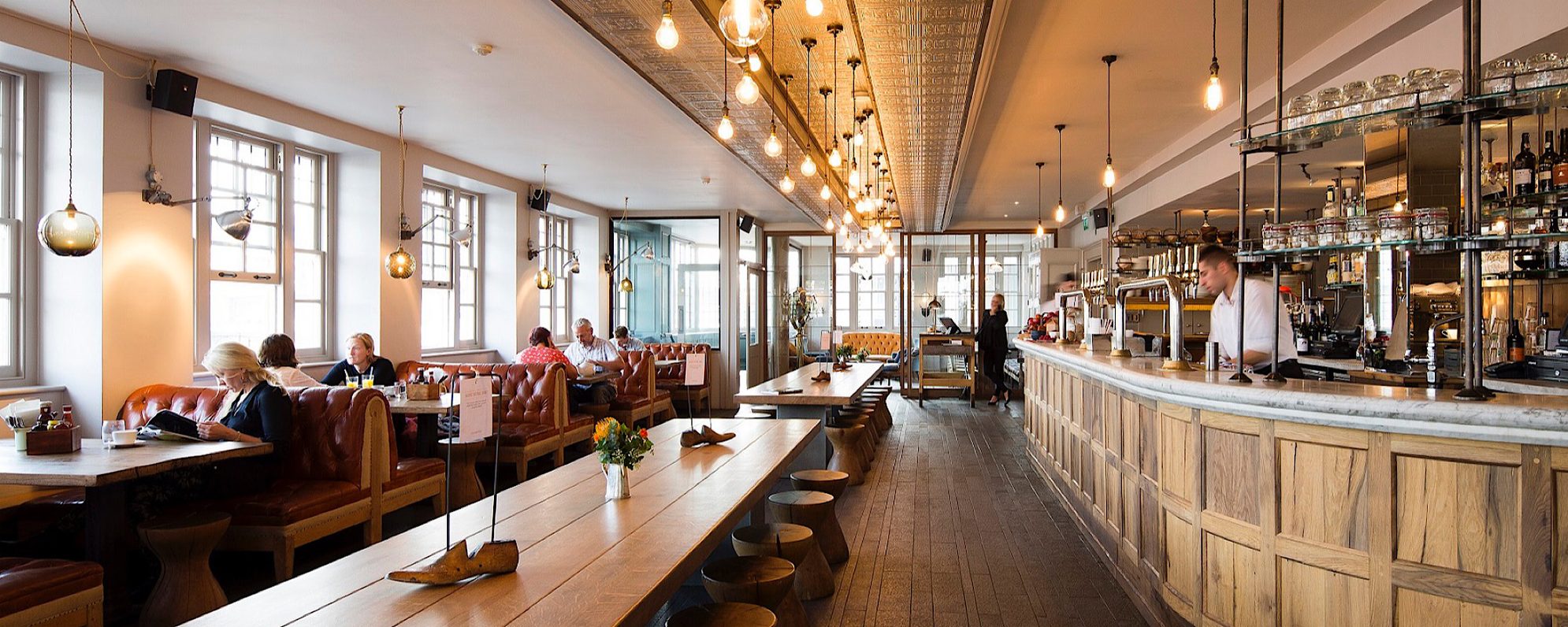 A bright and open bar space has bench and booth seating, lit by large bulbs from the ceiling