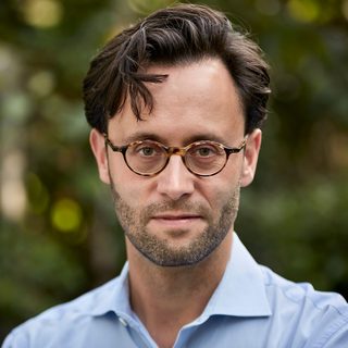 A person with dark hair, brown eyes and tortoiseshell glasses looking into camera