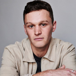 An actor with short brown hair wearing a cream-coloured shirt