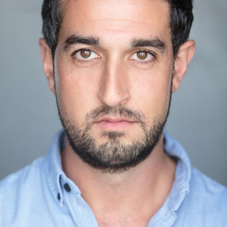 An actor looks into the camera. He has short dark hair and is wearing a blue shirt.