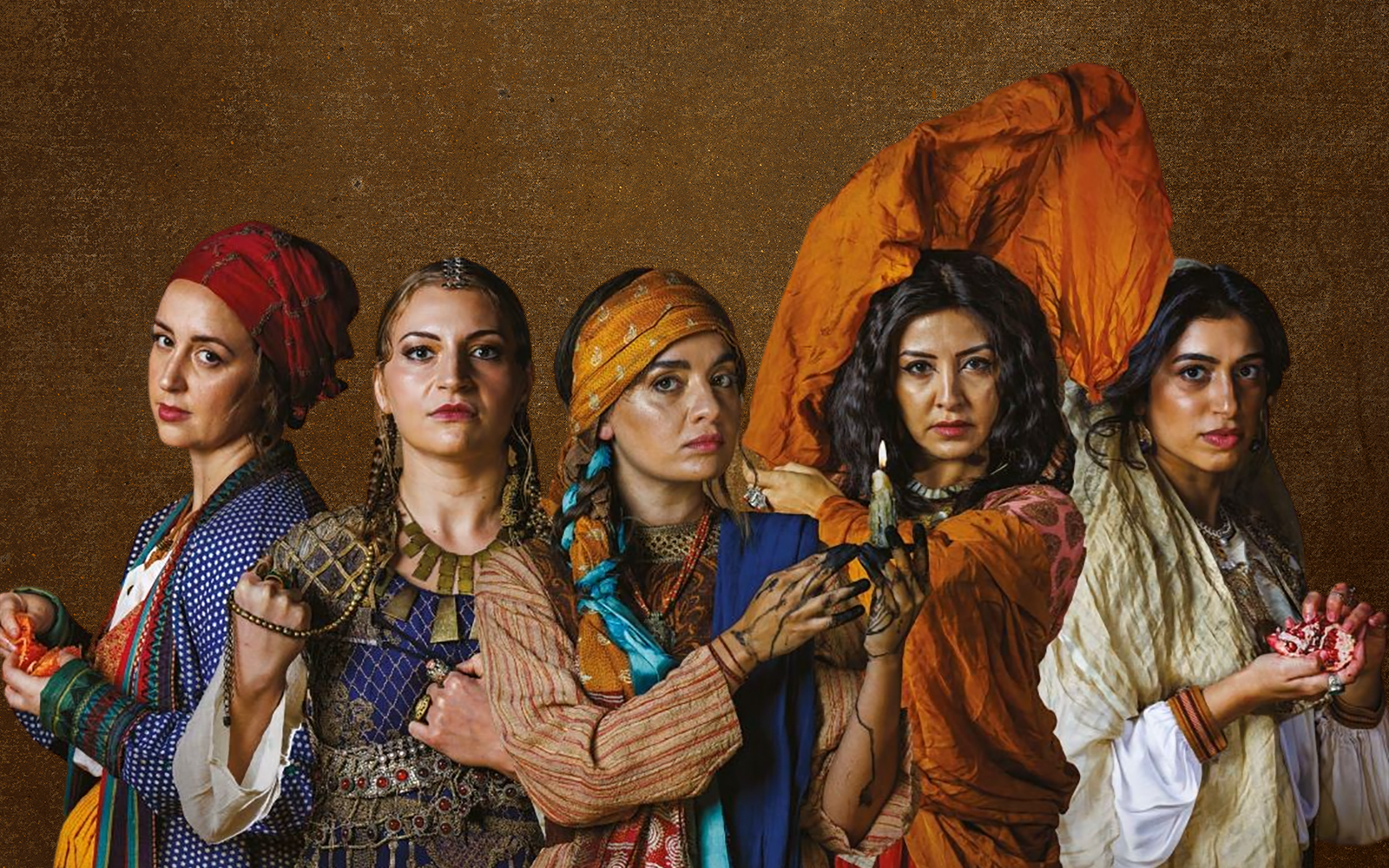 Five women look into the camera. They wear brightly coloured tunics and scarves, each holding a unique item. From left to right: a torn orange, a chain of beads, a candle dripping ink, a rust orange scarf in the air, and a crushed pomegranate.