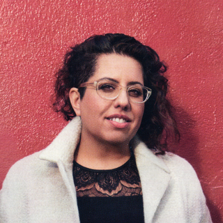 Headshot of Tina Gharavi wearing a black lace embellished top, white coat and clear glasses.